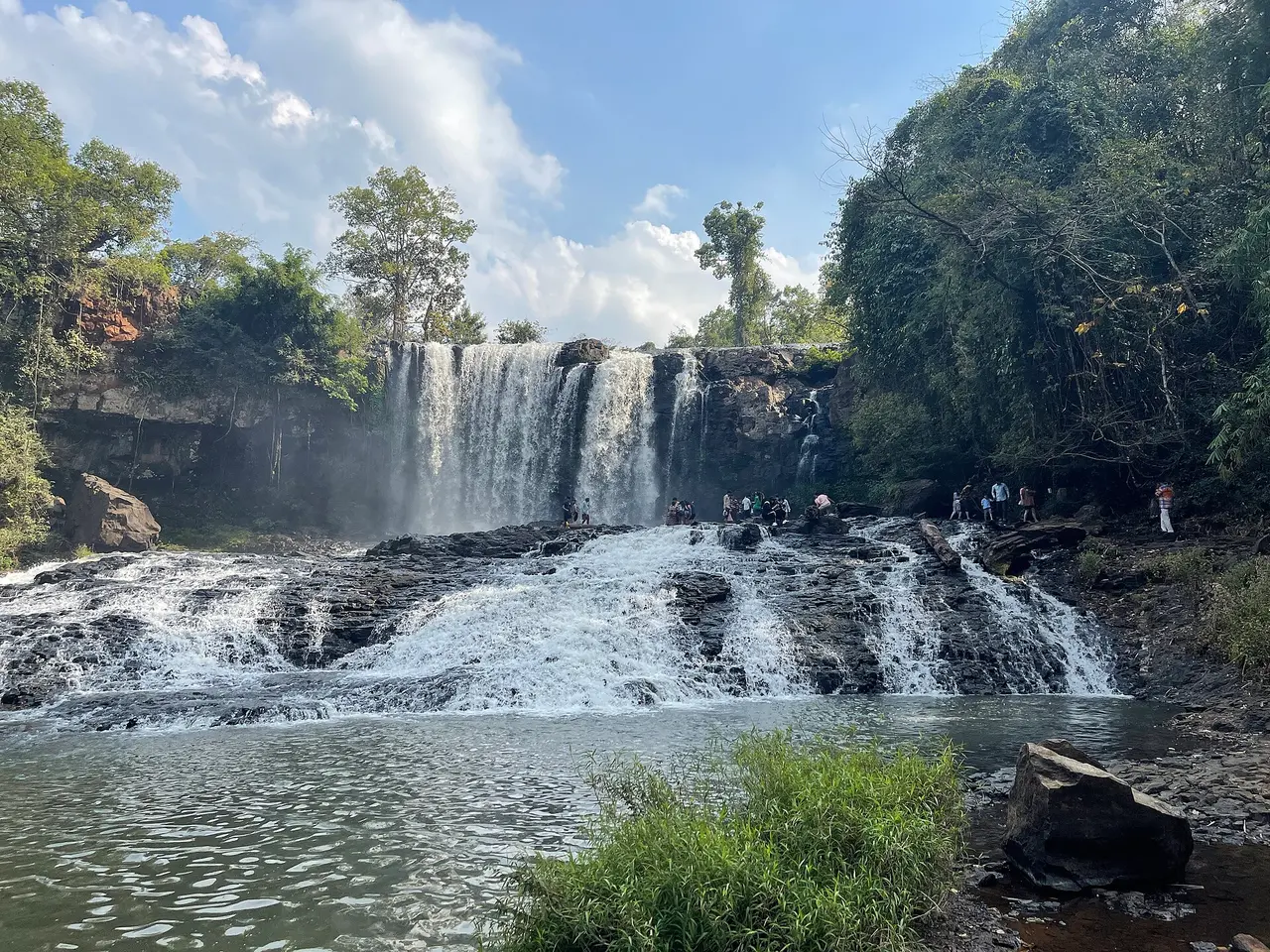 A wide two-tier waterfall pouring over a dark rock ledge into a pool, framed by forest