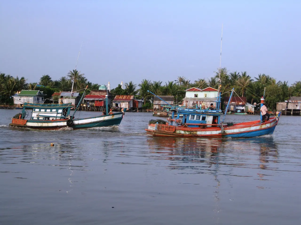 Painted wooden fishing boats moored on a wide river lined with palms and low buildings