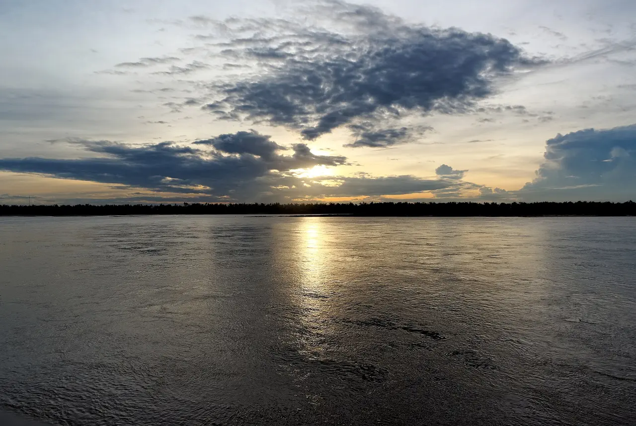 The sun setting behind clouds over a very wide, calm river
