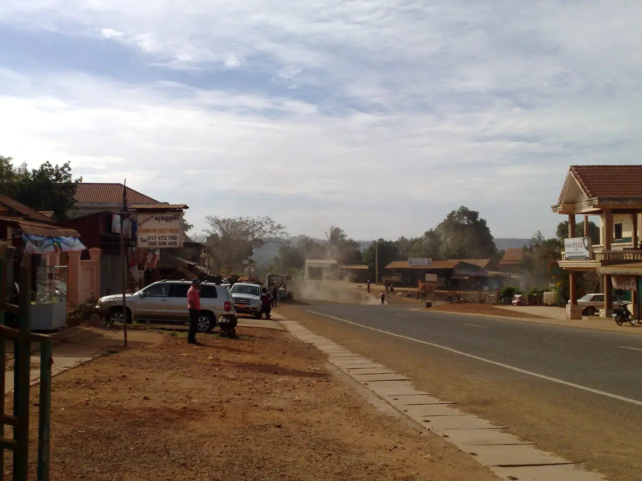 The wide main road of a small town with shophouses, parked cars and red dust